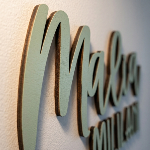 Custom wooden name wall sign featuring the name 'Malia' in a light sage green script with natural wood edges, mounted on a textured off-white wall.