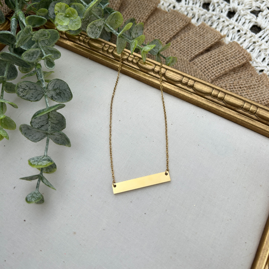 Gold necklace on a white surface with green leaves and a gold frame in the background