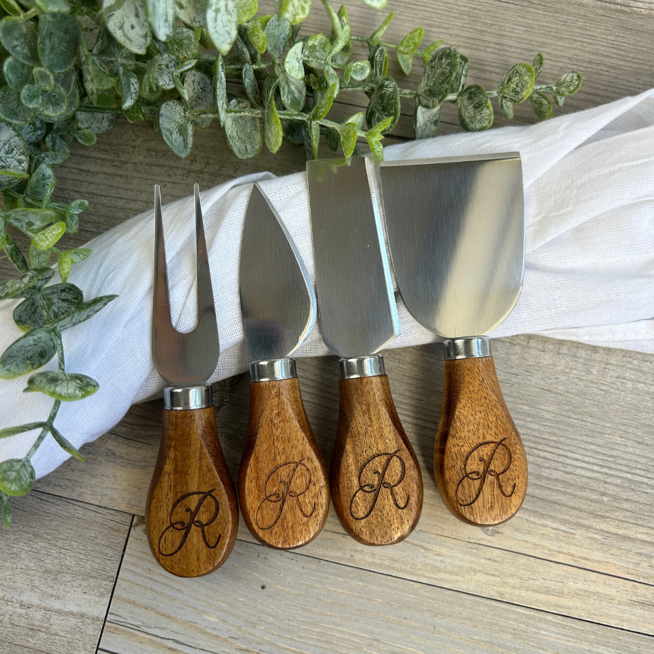 Four cheese knives with wooden handles and engraved initials on a wooden surface with a white cloth and greenery in the background.