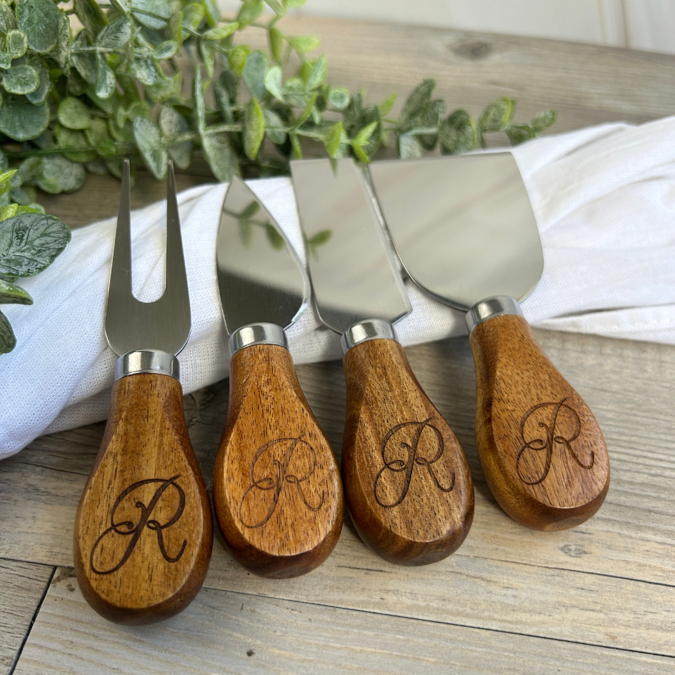 Set of four wooden-handled cheese knives with engraved initials on a wooden surface.
