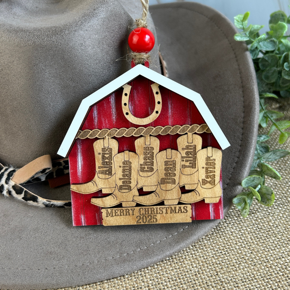 Wooden Christmas ornament shaped like a barn with engraved names and 'Merry Christmas 2025' text, hanging from a hat.