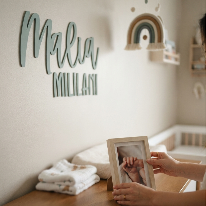 A personalized nursery wall sign reading 'Malia MILILANI' in a muted sage green color, mounted on a cream-colored wall in a softly lit baby nursery. In the foreground, hands are holding a small wooden picture frame on a wooden dresser next to folded white baby blankets.