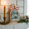 A decorative wooden picture frame ornament with engraved holiday motifs and a red-and-white striped bow, displayed on a white brick surface beside a lit candle, greenery, and cotton bolls.