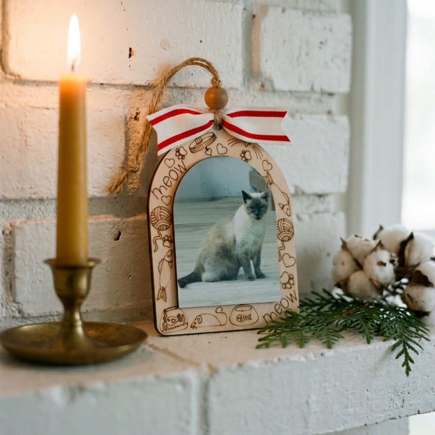 A decorative wooden picture frame ornament with engraved holiday motifs and a red-and-white striped bow, displayed on a white brick surface beside a lit candle, greenery, and cotton bolls.