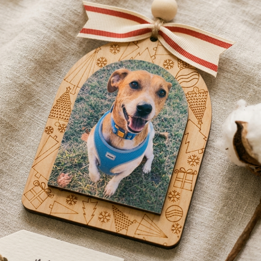 A personalized wooden Christmas ornament featuring a photo of a dog wearing a blue harness, hanging from a cream ribbon with red stripes, displayed on a textured beige fabric background.