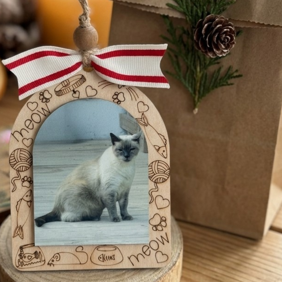 A wooden arch-shaped photo frame ornament with engraved cat-themed doodles and the word 'meow' on a ribbon, holding a photo of a cat, styled on a wooden surface with a brown gift bag and pine branch in the background.