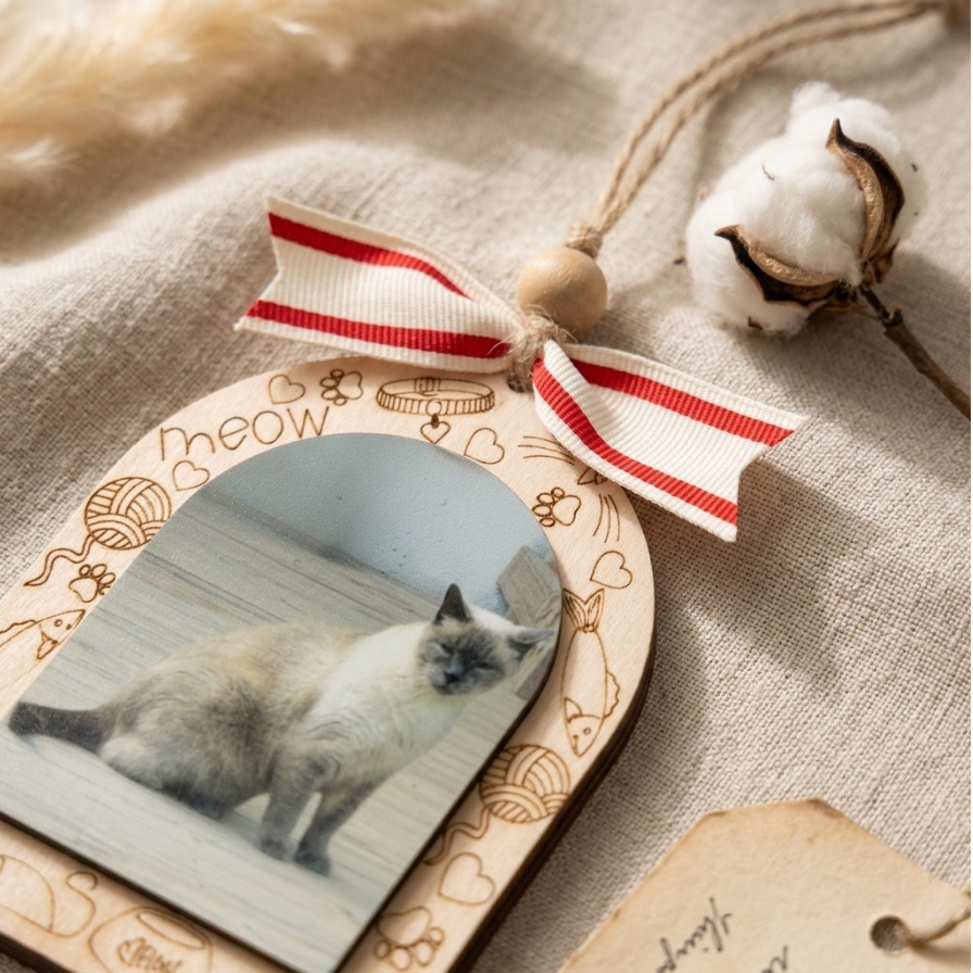 A wooden keepsake photo plaque with a cat photo in the center, decorated with a red and white striped ribbon bow and engraved cat illustrations. The plaque is styled on a beige linen fabric next to a cotton boll.