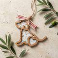 A wooden dog-shaped hanging ornament with a red and white striped ribbon, shown on a light stone surface with olive branches around it. The ornament has laser-engraved details including the words 'dog', paw prints, baseballs, and the name 'Lucy'.