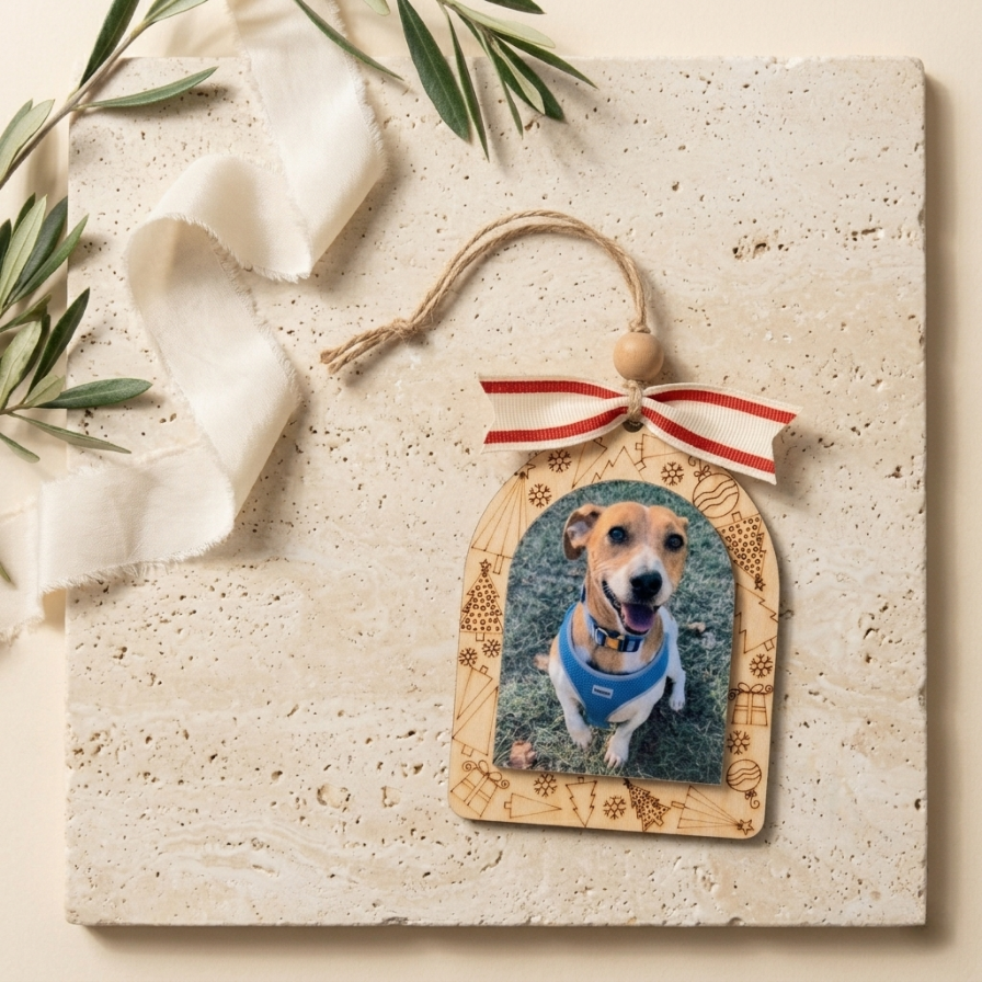 A personalized wooden hanging ornament featuring a photo of a dog wearing a blue harness, displayed on a textured beige stone tile with a cream ribbon and green leaves.