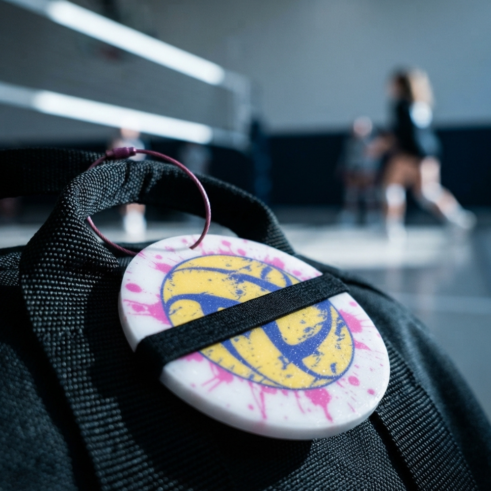 A round white and yellow volleyball-themed keychain or bag charm with pink paint splatter details, attached to a black strap on a black bag, with a blurred indoor sports court in the background.