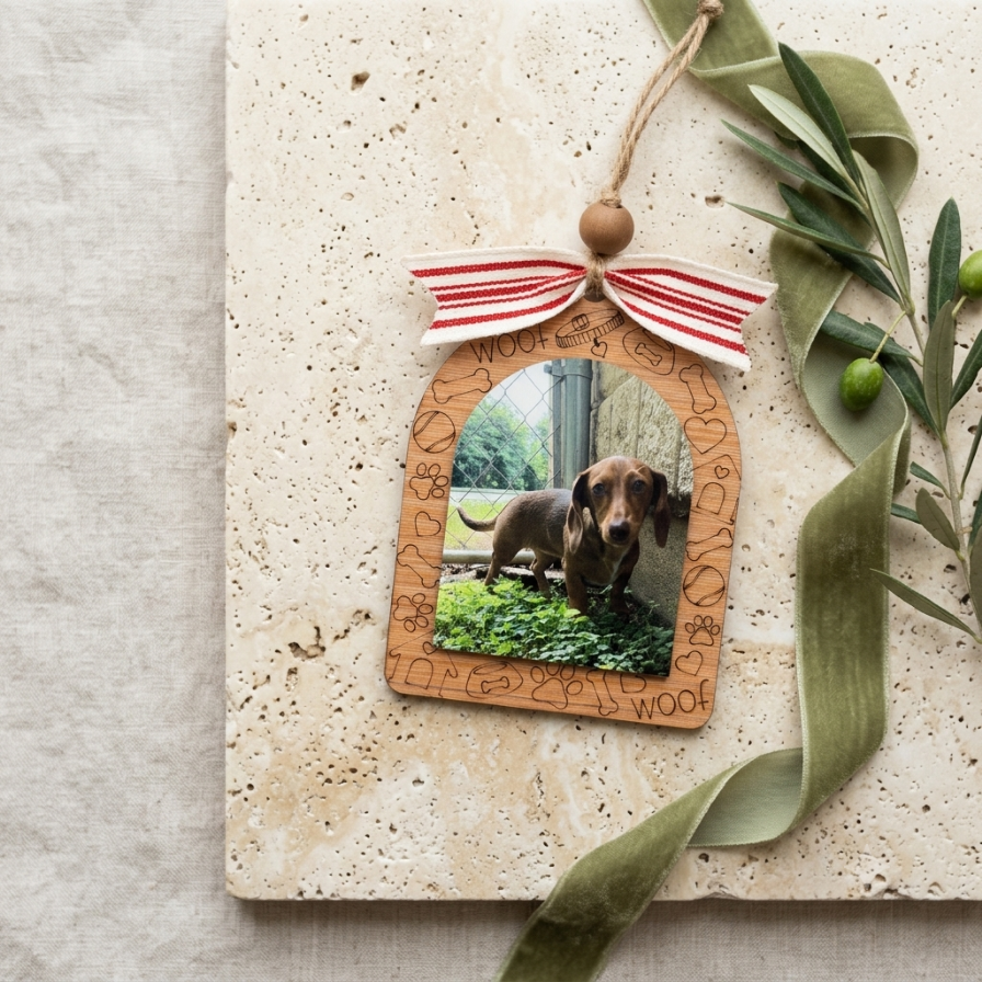 A wooden arch-shaped hanging ornament with a photo insert of a dachshund, decorated with a red-and-white striped ribbon and a wooden bead at the top, styled on a stone board with green ribbon and olive branches.