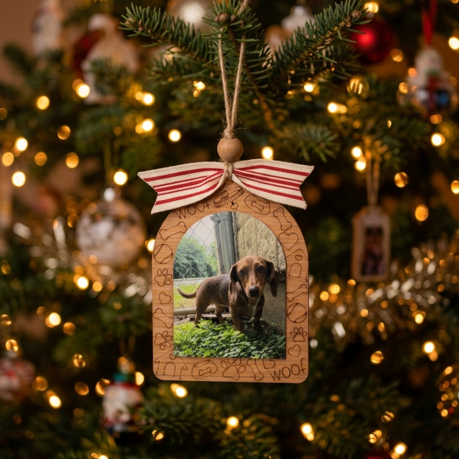 A wooden arched Christmas ornament hanging on a decorated Christmas tree. The ornament features a photo of a dachshund dog in the center, with engraved dog-themed phrases around the border, a red and white striped ribbon bow at the top, and a wooden hanging loop.