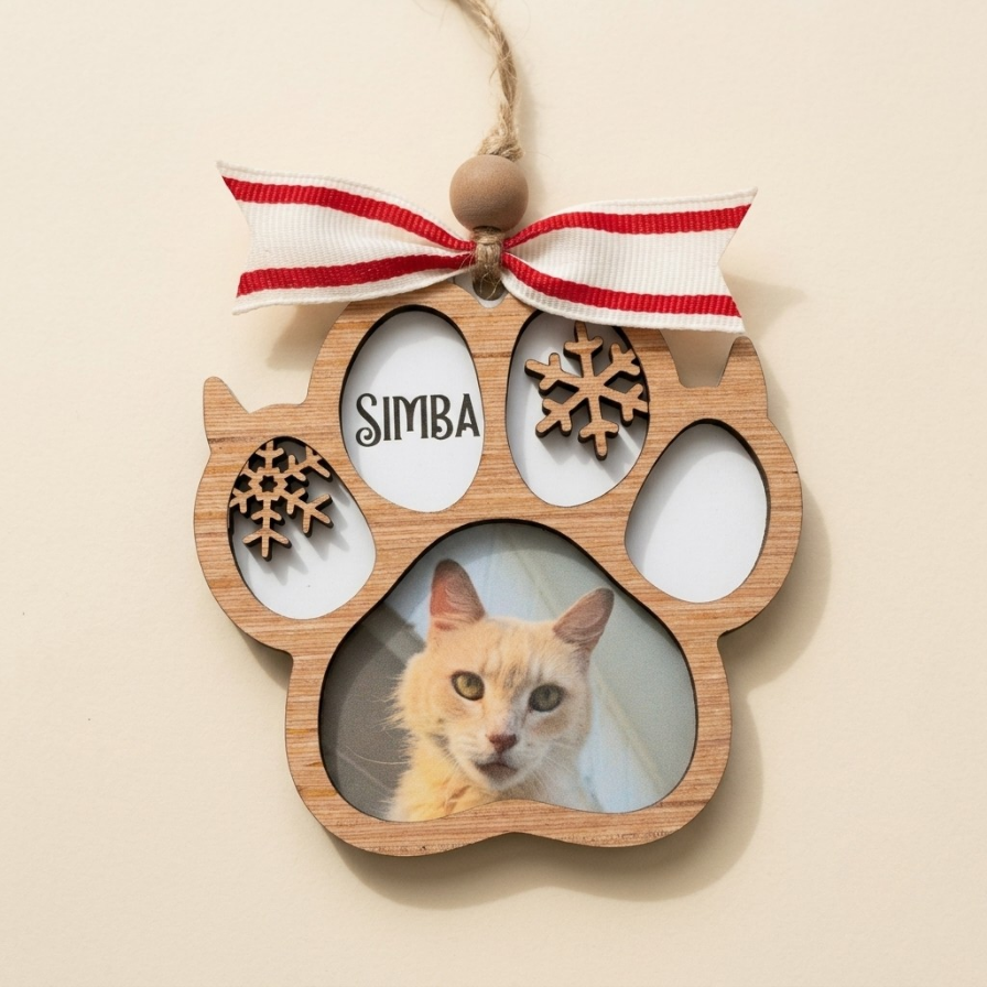 A wooden paw-shaped hanging ornament with a red and white striped ribbon at the top, featuring cutout snowflakes and the name 'SIMBA' in one eye slot, with a photo of an orange cat in the paw pad slot, shown on a plain beige background.