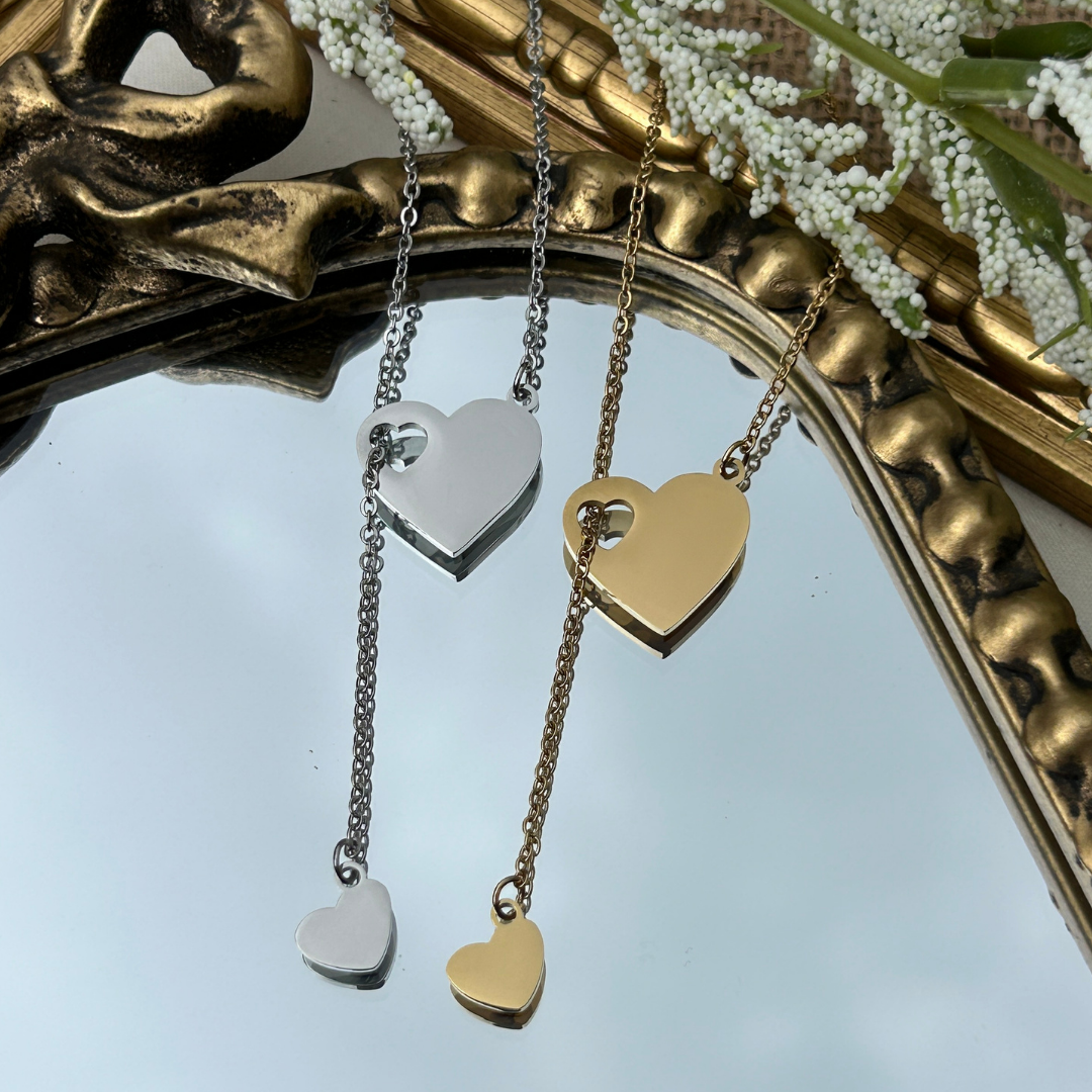 Two heart-shaped pendants, one silver and one gold, hanging from chains against a decorative mirror background.