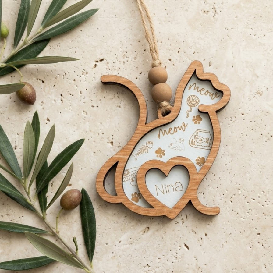 A wooden cat-shaped hanging ornament with a heart-shaped cutout in the center, engraved with the words 'meow' and 'Nina' along with small paw-print and yarn icons. The ornament is styled on a textured beige surface with olive branches and acorns.