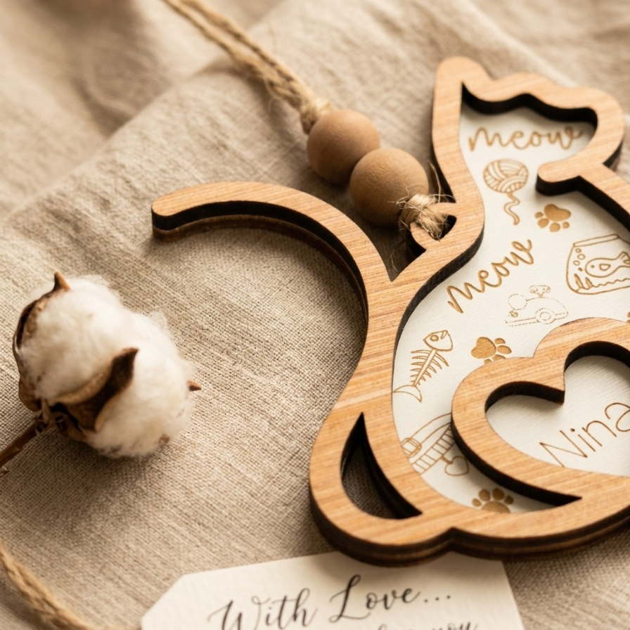 A wooden cat-shaped hanging ornament with layered cutout sections, bead string, and printed cat-themed illustrations and text, styled on beige fabric beside a cotton boll.