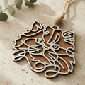 A laser-cut wooden cat-shaped ornament with a natural wood grain finish and a hanging loop, displayed on a textured beige surface with eucalyptus leaves.