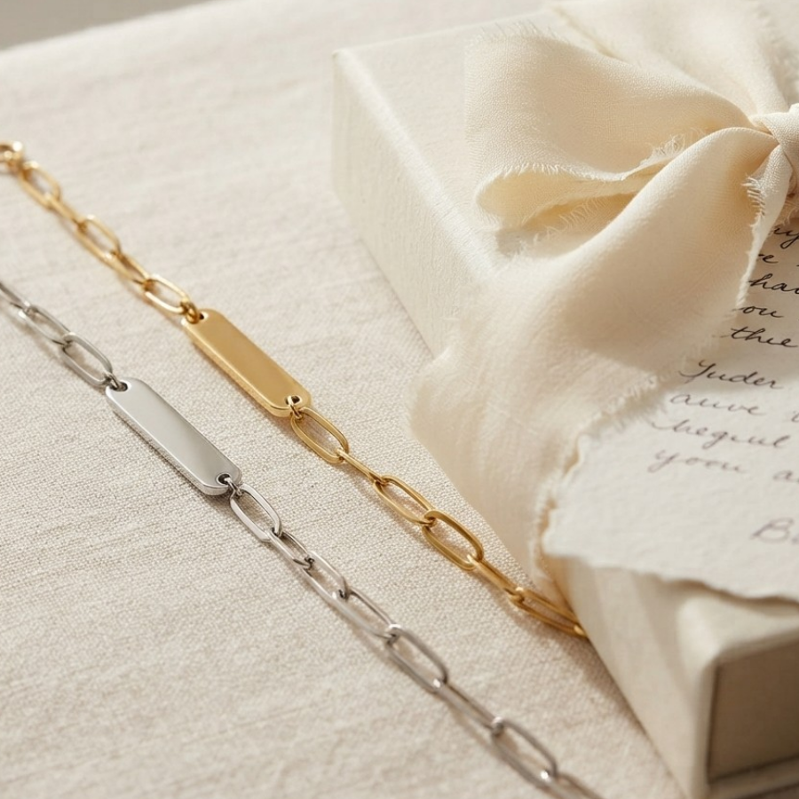 Two blank nameplate chain bracelets, one gold and one silver, displayed on a textured cream fabric next to a cream gift box with a satin bow.