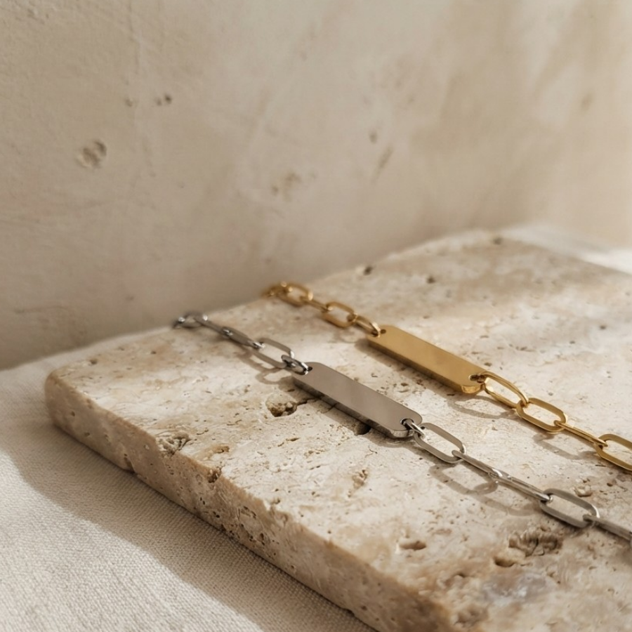 Two minimalist chain bracelets with blank rectangular nameplates, one in gold and one in silver, displayed on a textured beige stone slab.