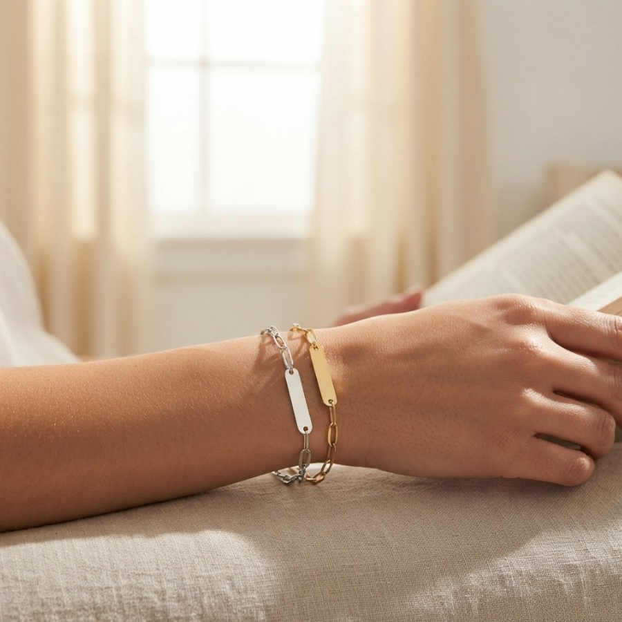 A close-up of a person's wrist wearing three delicate chain bracelets in silver, gold, and a two-tone silver-and-gold design, while holding an open book in a softly lit room.