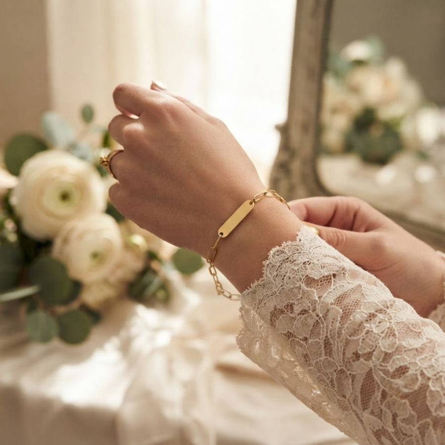 A delicate gold bar bracelet with a small chain link detail is shown being worn on a person's wrist, paired with a matching gold ring. The person is wearing a cream lace long-sleeve top, and the background features a soft-focus mirror and white flowers.