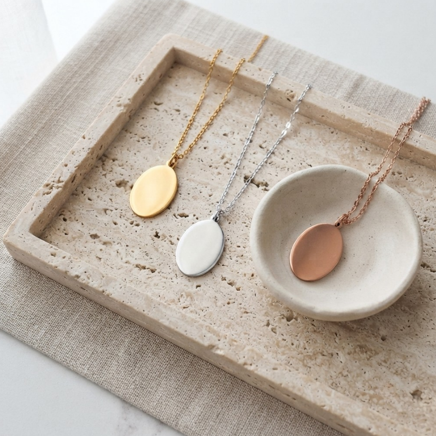 Three minimalist oval pendant necklaces in gold, silver, and rose gold, displayed on a textured beige linen cloth and a small ceramic dish.