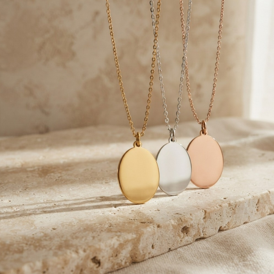 Three oval pendant necklaces in gold, silver, and rose gold, hanging on delicate chains against a warm beige stone background.