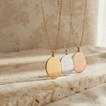 Three oval pendant necklaces in gold, silver, and rose gold, hanging on delicate chains against a warm beige stone background.