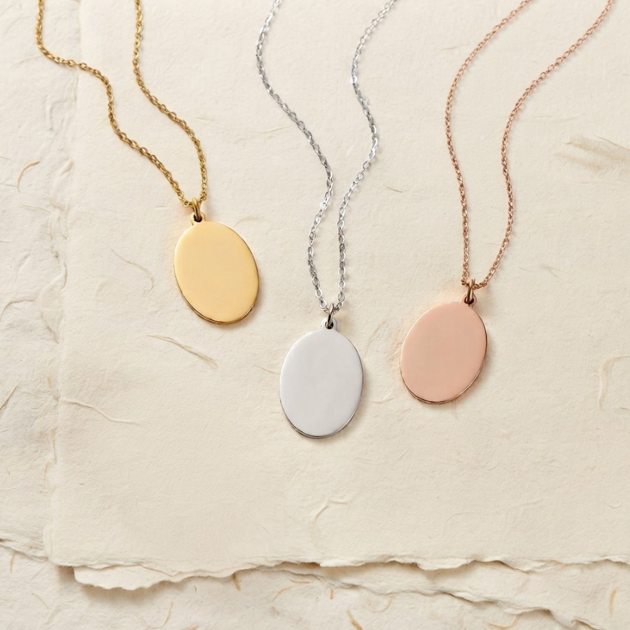 Three oval pendant necklaces on delicate chains, displayed on a textured beige paper background. The necklaces feature gold, silver, and rose gold pendants.