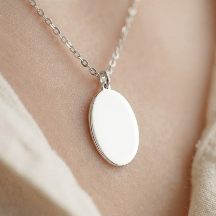A close-up of a silver oval pendant necklace worn on a person's neck, with a plain white face on the pendant and a soft beige top in the background.