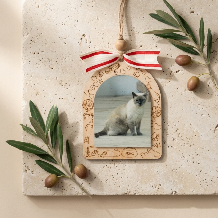 A decorative wooden arch-shaped hanging ornament with a photo slot, shown on a beige stone surface with olive branches and olives. The ornament has engraved cat-themed doodles and a red-and-white striped ribbon bow at the top.