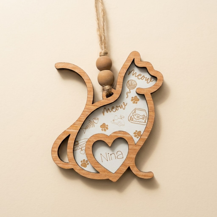 A wooden cat-shaped hanging ornament with a natural light wood finish, featuring a heart-shaped cutout with the name 'Nina' and paw prints, suspended by a twine string with three wooden beads against a warm beige background.
