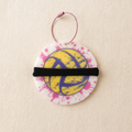 A round white disc ornament featuring a yellow and purple volleyball graphic, decorated with pink paint splatters and a black elastic band across the center, hanging from a pink cord on a textured beige background.