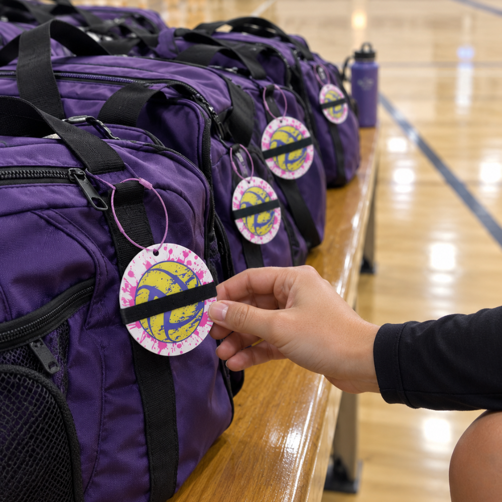 A close-up of a purple athletic duffel bag on a wooden bench in a gym, with a person's hand adjusting the black strap attached to a round branded tag.