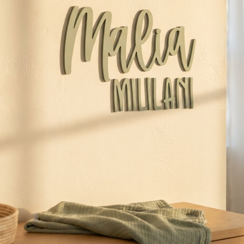 Personalized wooden name wall sign reading 'Malia MILANI' in a light sage green color, mounted on a textured beige wall above a wooden table with a folded green cloth.